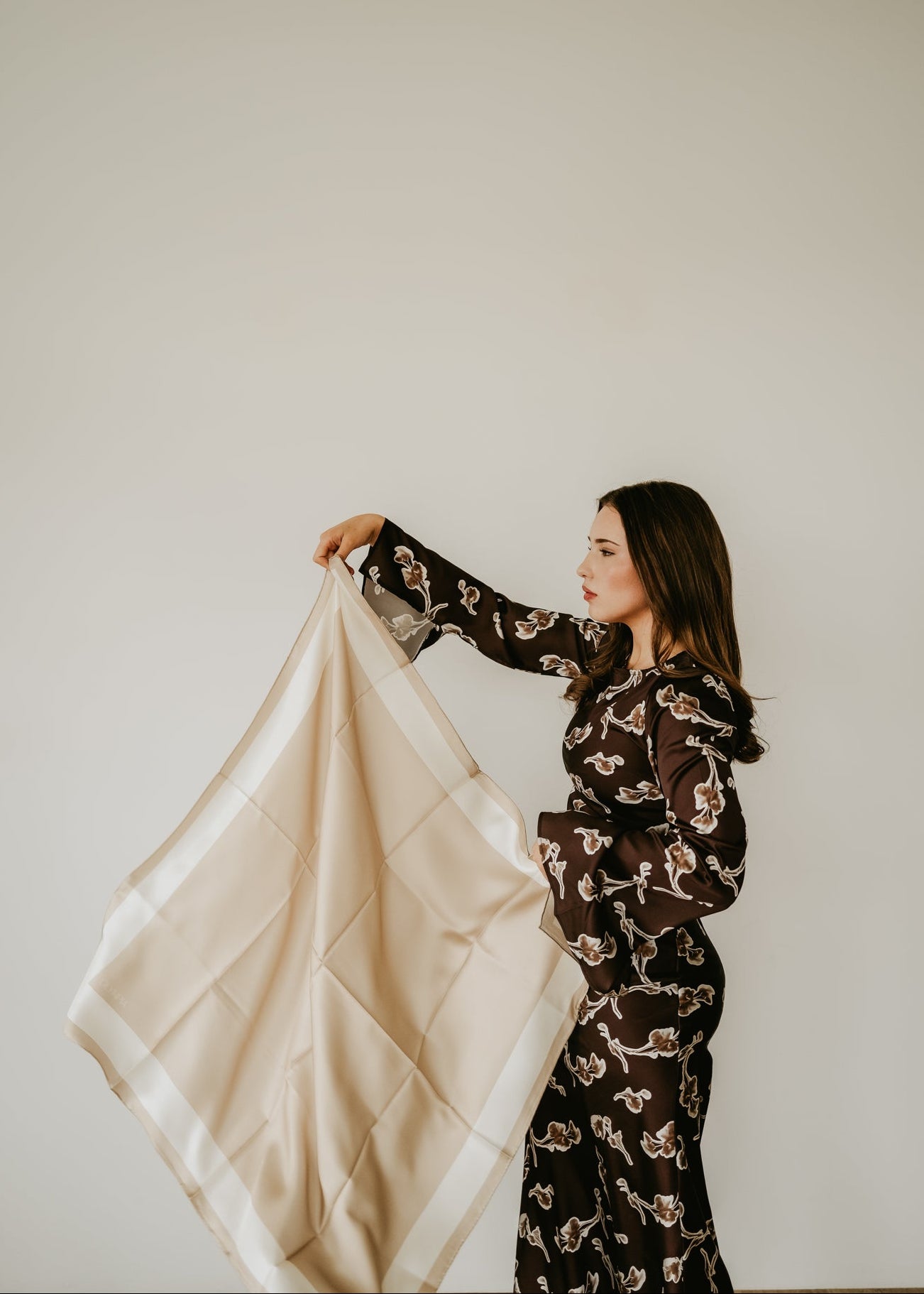 Woman holding a beige silk scarf against a plain wall
